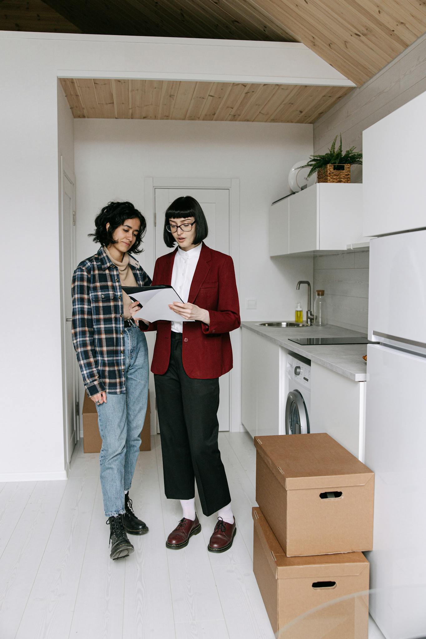 Two women discuss paperwork in a modern kitchen with moving boxes around them, symbolizing a new home purchase.